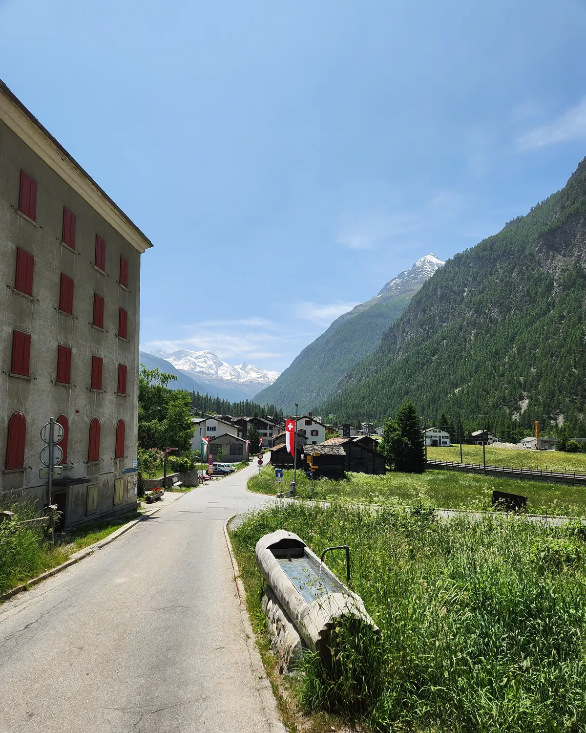 Mountain water fountain in Randa village with alpine houses, meadow and snowy peaks in the background.