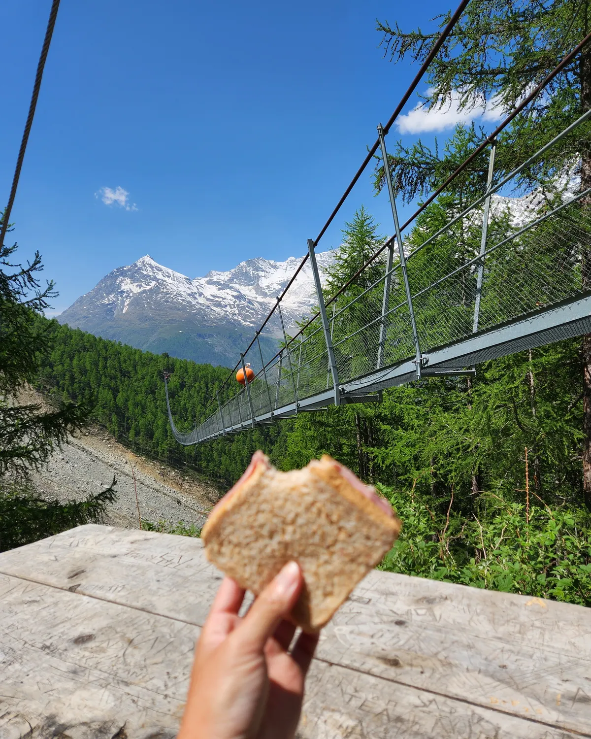 Hand holding a sandwich beside the Charles Kuonen Suspension Bridge with snowy mountains in the background.