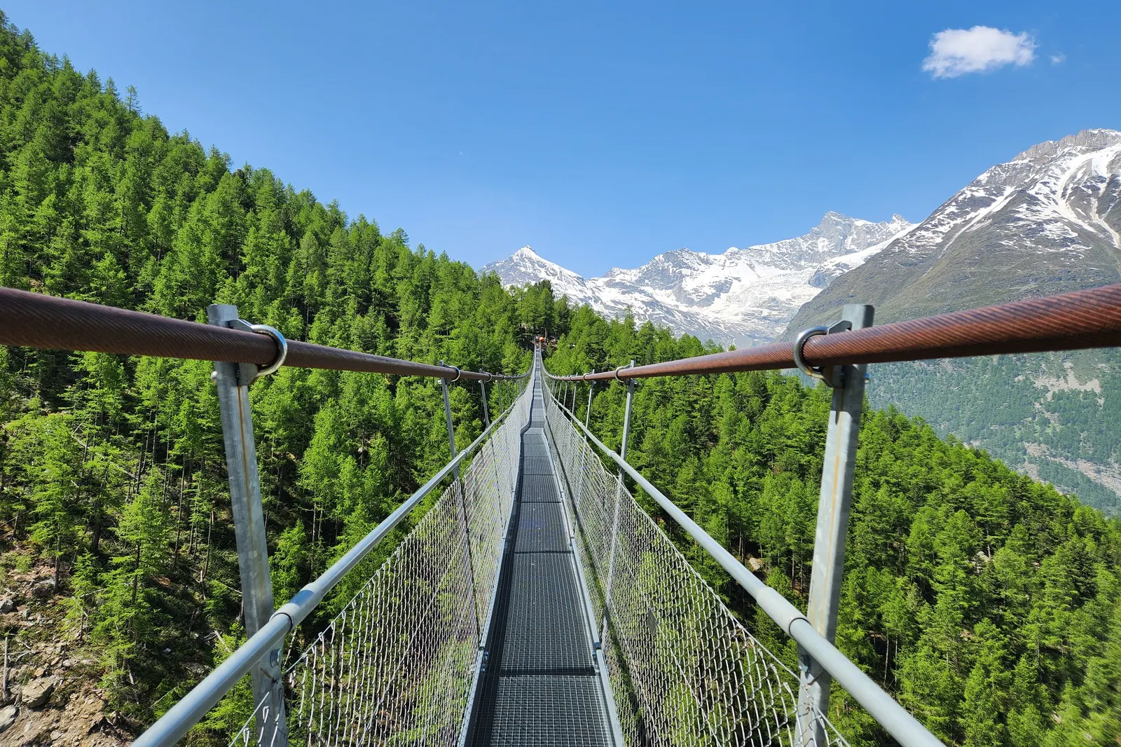 Charles Kuonen Suspension Bridge stretching across a green alpine valley near Randa, with snowy mountains behind.